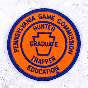 Peut inclure: Un écusson brodé circulaire avec un fond orange et une bordure bleue. Le texte indique PENNSYLVANIA GAME COMMISSION, HUNTER, GRADUATE, TRAPPER et EDUCATION. Un contour bleu en forme de clé de voûte est centré sur l'écusson.