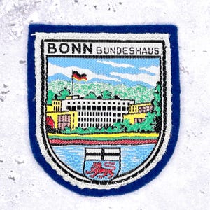 May include: Embroidered patch featuring the text "BONN BUNDESHAUS" above a detailed image of a building, a river, and a coat of arms. The patch has a blue border and a white stitched edge. A German flag is visible.