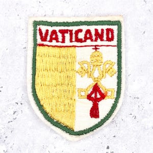 May include: A shield-shaped embroidered patch featuring the word VATICAND in red thread at the top. The design includes a yellow vertical section on the left and a gold-colored emblem of crossed keys with a red cord on the right, bordered in green.