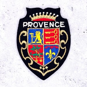 May include: Embroidered patch with a black border and a heraldic design. The patch features the word "PROVENCE" above a shield divided into four sections with various symbols, including keys and a fleur-de-lis. A gold crown sits at the top.
