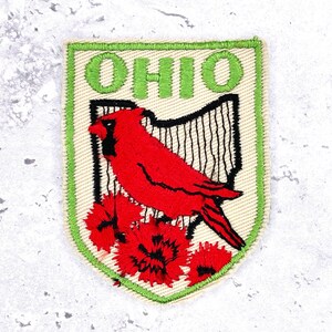 May include: A shield-shaped embroidered patch with a beige background and a green border. It features the word OHIO in green letters above a black outline of the state, a red cardinal bird, and three red carnation flowers at the bottom.