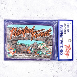 May include: A rectangular woven patch featuring the words "Petrified Forest" in orange script over a landscape scene. The patch includes a blue sky, gray rock formations, and the text "National Park, Arizona". The patch is packaged in a clear plastic bag.