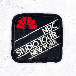 Puede incluir: Un parche bordado cuadrado con fondo negro y un borde negro grueso. El parche presenta un logotipo de pavo real rojo estilizado en la esquina superior izquierda y texto blanco en diagonal que dice NBC STUDIO TOUR NEW YORK en tres líneas.