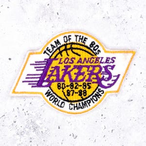 May include: Embroidered patch featuring the Los Angeles Lakers logo in purple and gold. The patch reads "TEAM OF THE 80s" and "WORLD CHAMPIONS" with the years 80-82-85 and 87-88.
