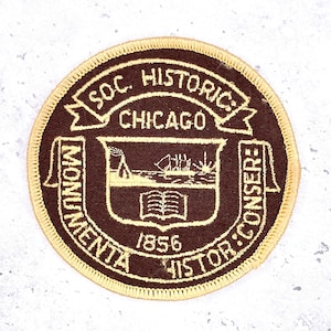 May include: A circular patch with a brown background and gold trim. The patch features the text "SOC. HISTORIC CHICAGO" and "MONUMENTA HISTOR: CONSER:" around the edge. A central emblem depicts a building, ship, and open book, with the year "1856".