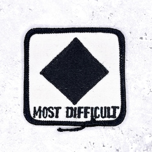 Op de afbeelding: Vierkantige patch met een zwarte ruit op een witte achtergrond, omlijnd door een zwarte rand. De woorden "MOST DIFFICULT" zijn eronder in het zwart geborduurd. Deze patch is waarschijnlijk voor skipistes of wandelpaden.