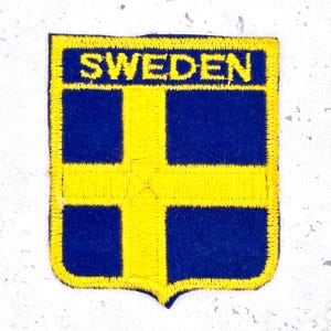 May include: Embroidered patch featuring the flag of Sweden. The patch has a blue background with a yellow Nordic cross. The word "SWEDEN" is embroidered in yellow at the top. The patch has a yellow border.