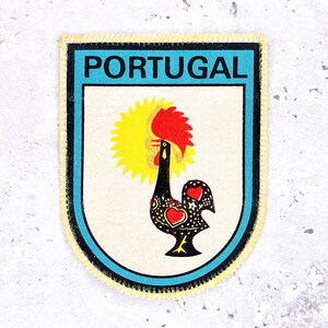 May include: A rectangular patch with a blue border and the word "PORTUGAL" in white letters. The patch features a stylized rooster with a yellow sunburst, red comb, and black body with heart and dot patterns.