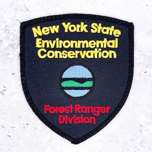 Op de afbeelding: Een schildvormige geborduurde patch met een zwarte achtergrond. De tekst New York State Environmental Conservation is in het geel geborduurd. Een centraal rond embleem toont lichtblauwe en groene lagen. Forest Ranger Division is in het rood.