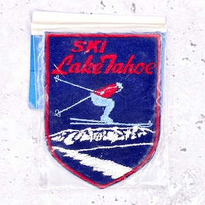 Pode incluir: Remendo bordado azul e vermelho com as palavras "SKI Lake Tahoe" e um esquiador. O remendo tem uma borda vermelha e uma cordilheira branca na parte inferior. Está em um saco plástico transparente.
