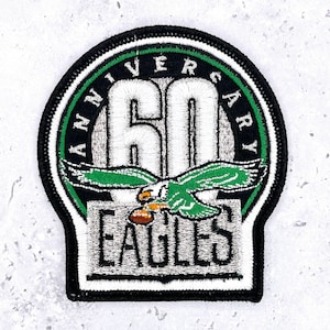 Op de afbeelding: Geborduurde patch met een zwarte rand, met een groen en wit ontwerp. De patch herdenkt het "60" jarig jubileum van het "EAGLES" team. Een groene adelaar met een voetbal staat in het midden, met het woord "ANNIVERSARY" erboven.