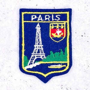 May include: A blue embroidered patch with a yellow border. The patch features the word "PARIS" at the top, a white Eiffel Tower, green trees, and a red and yellow crest. The patch is shield-shaped.