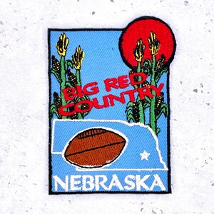 Op de afbeelding: Geborduurde patch met een blauwe achtergrond, met de woorden "BIG RED COUNTRY" in het rood. De patch bevat een voetbal, maïskolven, een rode zon en de omtrek van Nebraska. Het woord "NEBRASKA" staat onderaan.