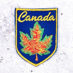 May include: Embroidered patch featuring the word "Canada" in yellow script above a colorful maple leaf. The leaf is red, yellow, and green, set against a blue background with a yellow border. The patch is shield-shaped.
