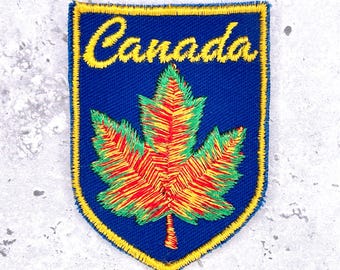 May include: Embroidered patch with the word "Canada" in yellow script above a colourful maple leaf. The leaf is red, yellow, and green, set against a blue background with a yellow border. The patch is shield-shaped.