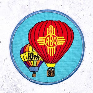 May include: Embroidered patch featuring two hot air balloons. The larger balloon is red with a yellow design and the letters "ABQ". The smaller balloon is multi-colored with the number "10th". The patch has a blue border.