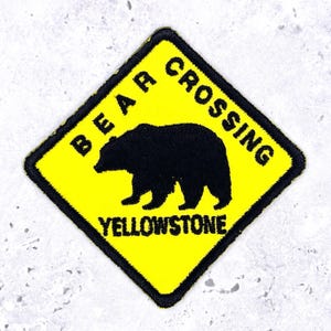 May include: A diamond-shaped embroidered patch with a bright yellow background and a black border. The patch features a black silhouette of a bear in the center, with the black text BEAR CROSSING arched above and YELLOWSTONE printed below.