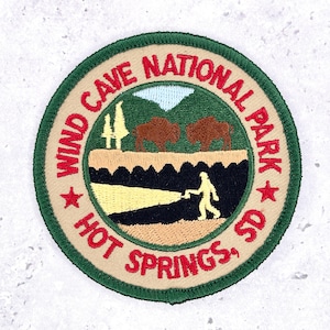 May include: A circular embroidered patch with a dark green border and tan background. It features red text reading WIND CAVE NATIONAL PARK and HOT SPRINGS, SD. The design shows two brown bison, green trees, a light blue sky, and a yellow silhouette figure.
