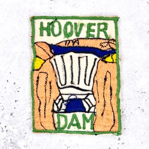 Può includere: Una toppa ricamata rettangolare con il testo HOOVER DAM in filo verde. Il design raffigura una struttura beige stilizzata con una sezione centrale bianca, accenti blu e riflessi gialli, il tutto bordato da un spesso bordo cucito verde.