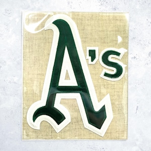May include: A rectangular patch featuring the dark green and white Oakland Athletics logo with the letter A and the letters 'S. The design is set against a textured, light tan background with a visible stitched border around the letters.