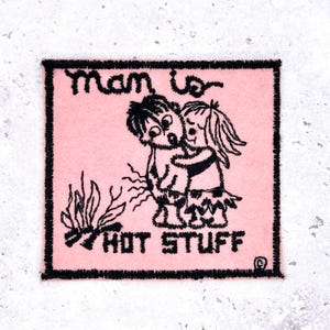 May include: A pink square patch with black embroidered text and design. The text reads "man is HOT STUFF" above a cartoon couple embracing. A small fire is depicted on the left side of the patch.