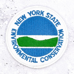 May include: A circular embroidered patch featuring a white border with the blue text NEW YORK STATE ENVIRONMENTAL CONSERVATION. The centre displays a blue sky above a white horizon line and a green rolling hill landscape over blue water.