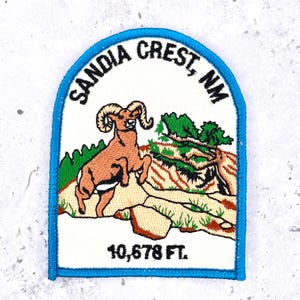 May include: An embroidered patch with a blue border featuring a bighorn sheep standing on a rocky landscape with green trees. The text reads SANDIA CREST, NM at the top and 10,678 FT. at the bottom, all set against a white background.