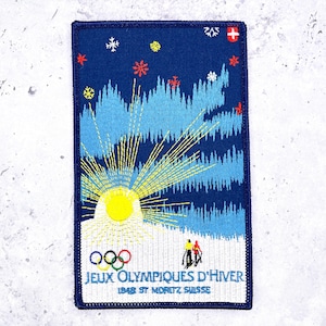 May include: A rectangular embroidered patch featuring a dark blue background with a yellow sun, light blue mountain silhouettes, and white snowflakes. It includes the Olympic rings, two skiers, and the text JEUX OLYMPIQUES D'HIVER 1948 ST MORITZ SUISSE.