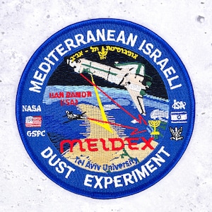 May include: A circular embroidered patch featuring a space shuttle over a map of the Mediterranean region. Text includes MEDITERRANEAN ISRAELI DUST EXPERIMENT, ILAN RAMON (ISA), NASA, GSFC, MELDEX, and Tel Aviv University, with flags and a menorah.