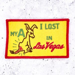 May include: A rectangular embroidered patch with a yellow background and a red border. It features a brown kangaroo illustration and green text that reads MY A I LOST IN, with the red cursive text Las Vegas below the kangaroo graphic.
