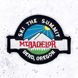 May include: A circular embroidered patch with a black border featuring a blue sky, a yellow sun, and a white mountain peak. The text reads "SKI THE SUMMIT" at the top, "MT. BACHELOR" in red across the center, and "BEND, OREGON" at the bottom.