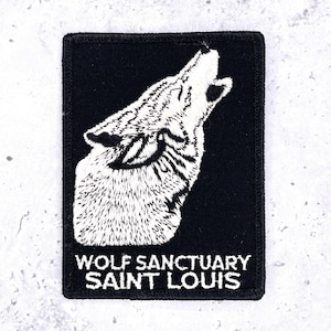 May include: Black rectangular patch featuring a white embroidered howling wolf. The text "WOLF SANCTUARY SAINT LOUIS" is embroidered in white below the wolf image. The patch has a black border and is likely made of fabric.
