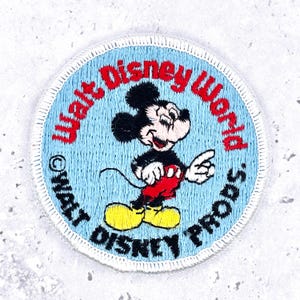 Puede incluir: Un parche bordado circular con fondo azul claro y una ilustración central de Mickey Mouse. El texto Walt Disney World está bordado en rojo, y el texto Walt Disney Prods. aparece en negro a lo largo del borde inferior del parche.