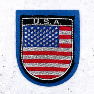 May include: A shield-shaped patch with a bright blue felt border. The center features an embroidered United States flag with silver stars and stripes. Above the flag, the text U.S.A. is stitched in silver thread on a black background.