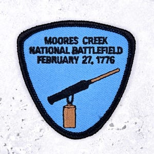 Può includere: Una toppa ricamata a forma di scudo con sfondo azzurro e bordo nero. Presenta il testo nero MOORES CREEK NATIONAL BATTLEFIELD FEBRUARY 27, 1776 sopra un grafico di un moschetto nero con calcio in legno marrone e bacchetta di caricamento.