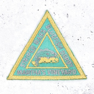 May include: A triangular embroidered patch with a teal center and a gold border. The patch features text that reads "MARTHA'S VINEYARD" and "SAVED BY THE SEA". A gray and gold image of a whale is in the center.