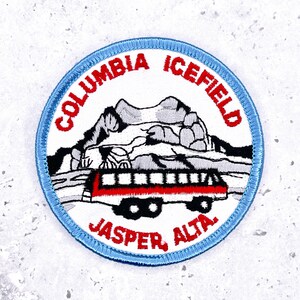 May include: Embroidered patch featuring a mountain scene and a red and black bus. The text "COLUMBIA ICEFIELD" and "JASPER, ALTA." are in red. The patch has a light blue border and is circular.