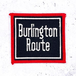 May include: Square patch with the words "Burlington Route" in white script on a black background. The patch has a white border and a red stitched edge.