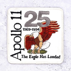 May include: A white embroidered patch commemorating the 25th anniversary of the Apollo 11 mission. The patch features a detailed eagle with red and brown wings, the text "Apollo 11", and the phrase "The Eagle Has Landed".