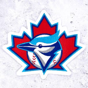 May include: An embroidered patch featuring a red maple leaf silhouette as the background. Centered on the leaf is a blue jay bird head profile in shades of blue, white, and grey, layered over a white baseball with red stitching details.