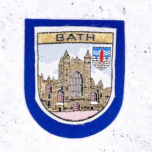 May include: A blue and white embroidered patch with the word "BATH" at the top. The patch depicts a detailed illustration of a cathedral and a small emblem, all within a blue shield-shaped border.