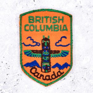 Könnte beinhalten: Orangefarbener, gestickter Aufnäher mit der Aufschrift "British Columbia Canada" und einem Totempfahl-Design. Der Aufnäher hat grüne, blaue und braune Details und ist schildförmig.