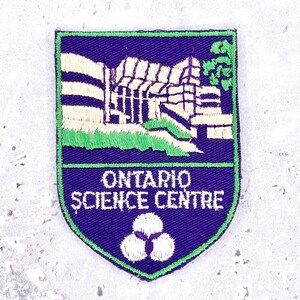 Pode incluir: Patch bordado em forma de escudo, apresentando uma ilustração de um edifício e o texto "ONTARIO SCIENCE CENTRE". O patch tem um fundo roxo com detalhes verdes e brancos e um emblema branco na parte inferior.