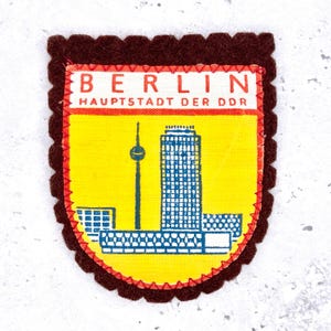 May include: A vintage Berlin souvenir patch. The patch features a yellow background with a blue and white illustration of a building and a tower. The text "BERLIN HAUPTSTADT DER DDR" is printed on a red banner. The patch has a brown border.