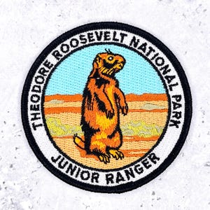May include: A circular embroidered patch featuring a brown prairie dog standing in a desert landscape with a light blue sky. The black border contains the white text Theodore Roosevelt National Park and Junior Ranger in a bold sans-serif font.