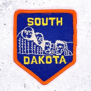 May include: A shield-shaped embroidered patch with a blue background and a bright orange border. It features white thread stitching depicting the Mount Rushmore monument and yellow embroidered text that reads SOUTH DAKOTA.