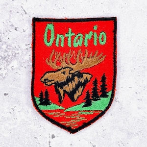 May include: Embroidered patch with a red background and a shield shape. The patch features the word "Ontario" in green, a brown moose, and green trees. The bottom of the patch has a green and red water design.