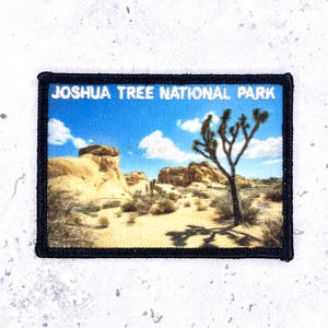 May include: A rectangular embroidered patch featuring a desert landscape with a Joshua tree, rock formations, and a blue sky with white clouds. The text JOSHUA TREE NATIONAL PARK is printed in white at the top. The patch has a thick black border.