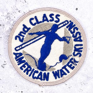 May include: Circular patch with embroidered text and a blue waterskier silhouette. The patch reads "2nd Class American Water Ski Assn.". The skier is in motion, holding a tow rope and skis. The patch has a cream-colored background.
