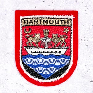 May include: A red patch with the word "DARTMOUTH" at the top. The patch features a crest with two lions, a boat, and wavy blue lines. The patch is bordered with white and black beads.